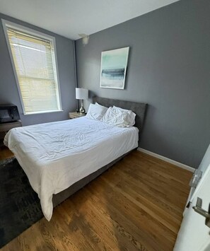 A bedroom with a bed, a nightstand, a lamp, a painting, and a window with blinds.