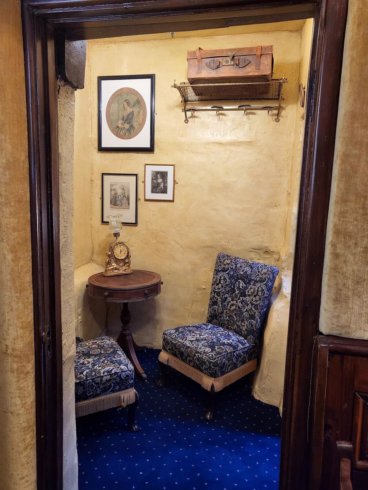A corner room with a patterned armchair, a small table with a clock, and a wall-mounted shelf with a leather bag.