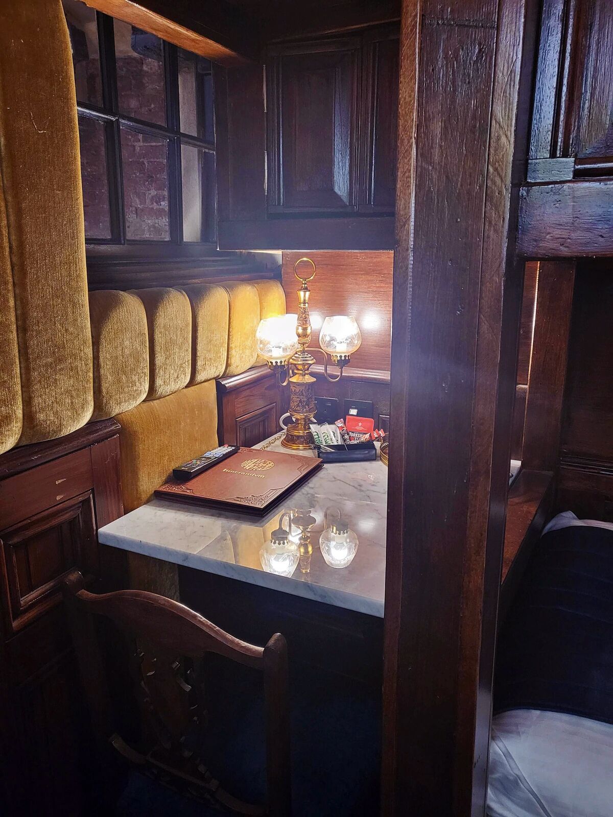 Un angolo accogliente con una panca imbottita, un tavolino in legno con un telecomando, una lampada decorativa e un piccolo vassoio con degli oggetti.