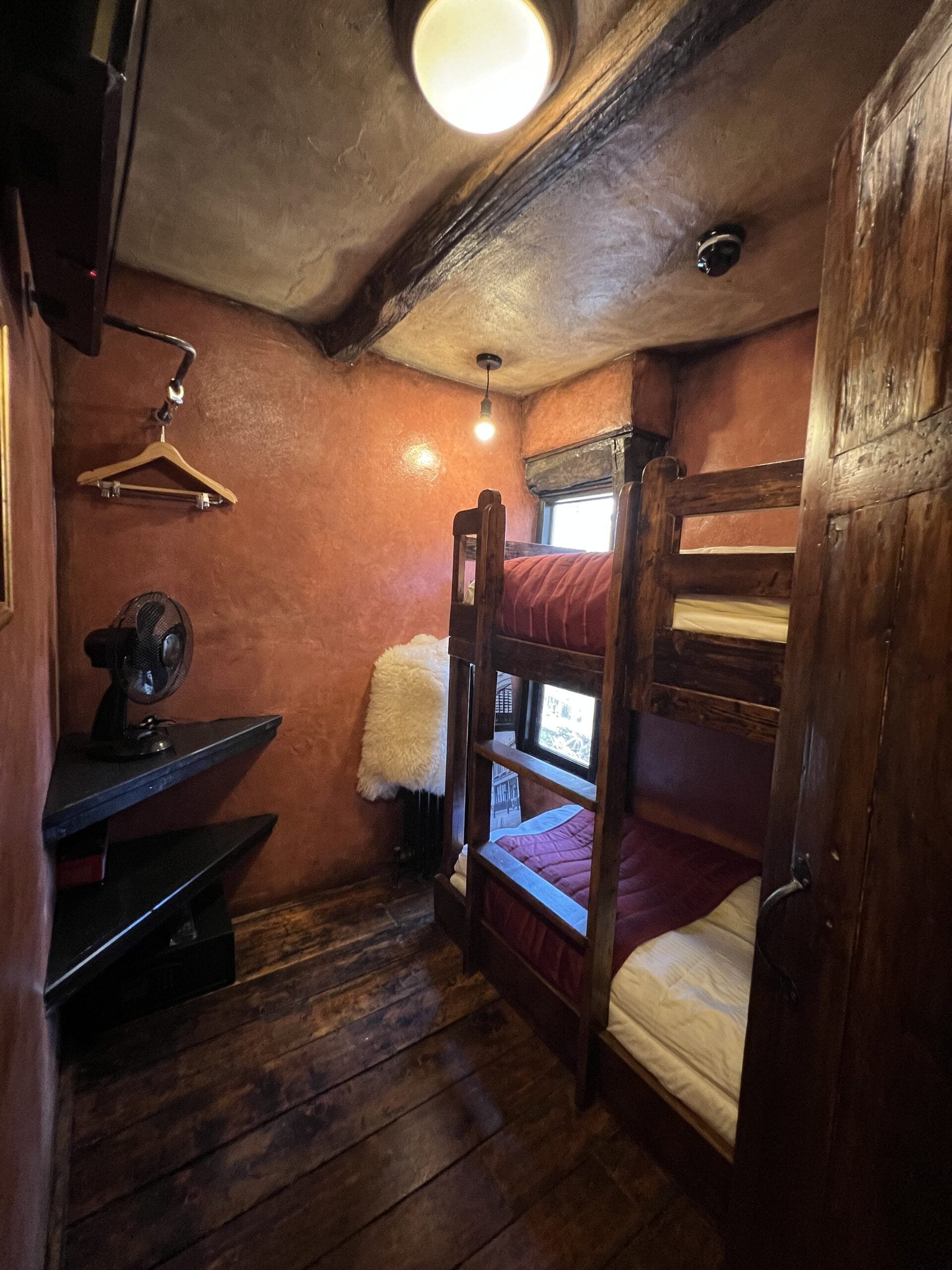 A rustic room with a bunk bed, a desk with a fan, a hanging light fixture, and a window with a view.