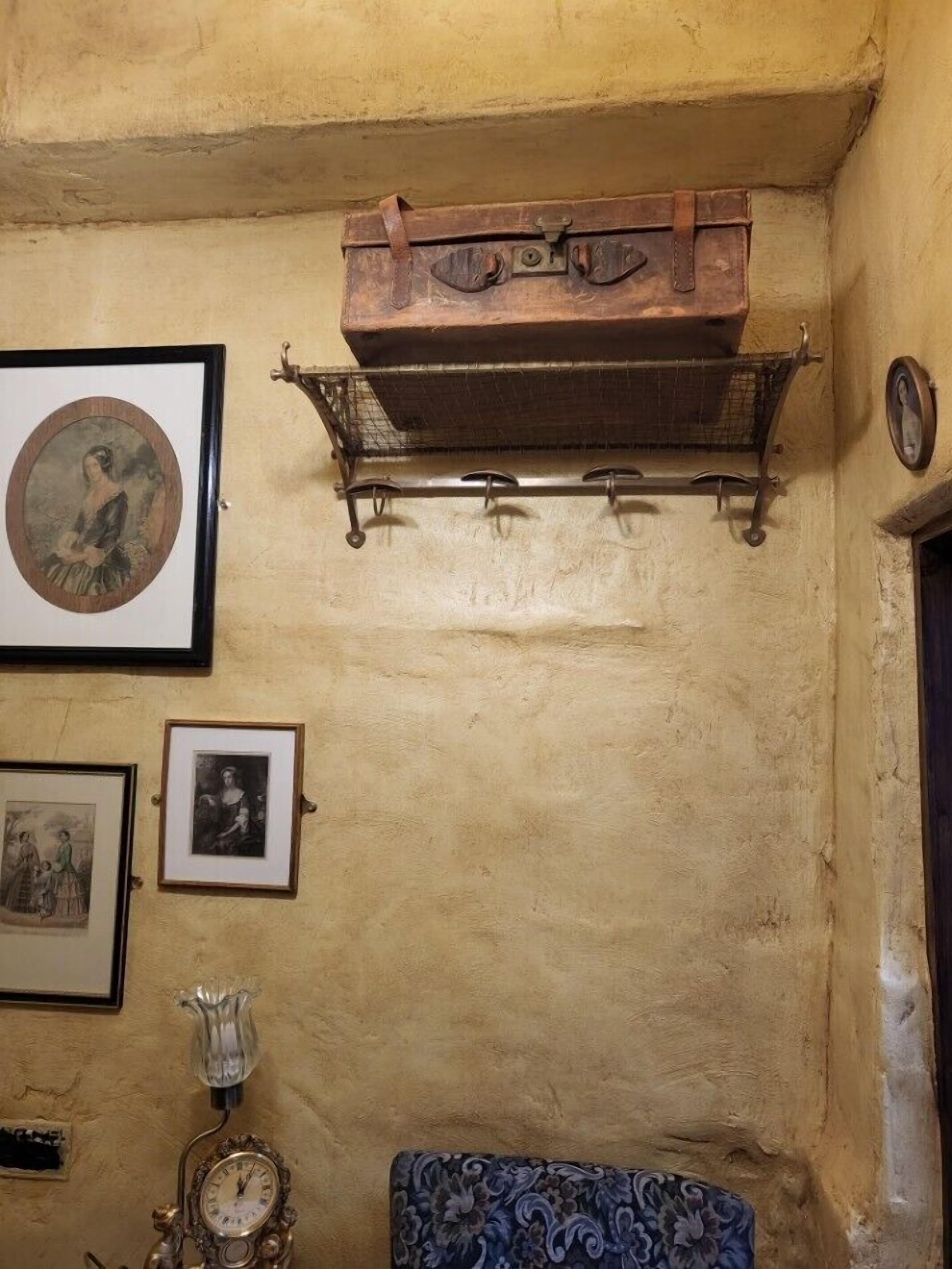 Una habitación con una maleta antigua, cuadros en la pared, un reloj y una silla estampada.