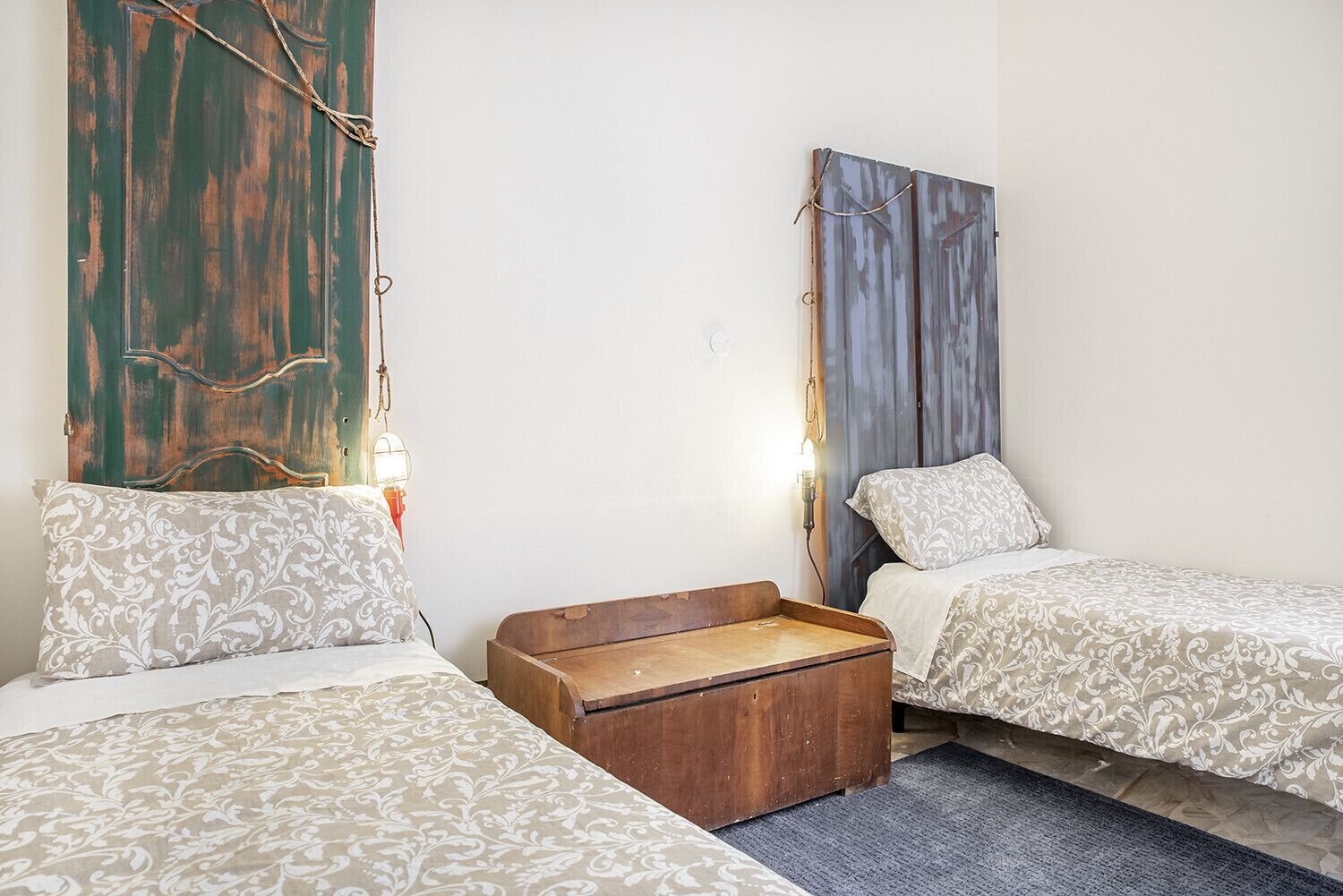 Een kleine, knusse kamer met twee bedden, een houten kist en een wandlamp.