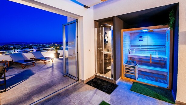 A modern hotel room with a glass door leading to a balcony with lounge chairs and a city view at dusk. - Penthouse Luxury Suite (Zadar)