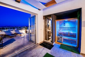 A modern hotel room with a glass door leading to a balcony with lounge chairs and a city view at dusk. - Penthouse Luxury Suite (Zadar)