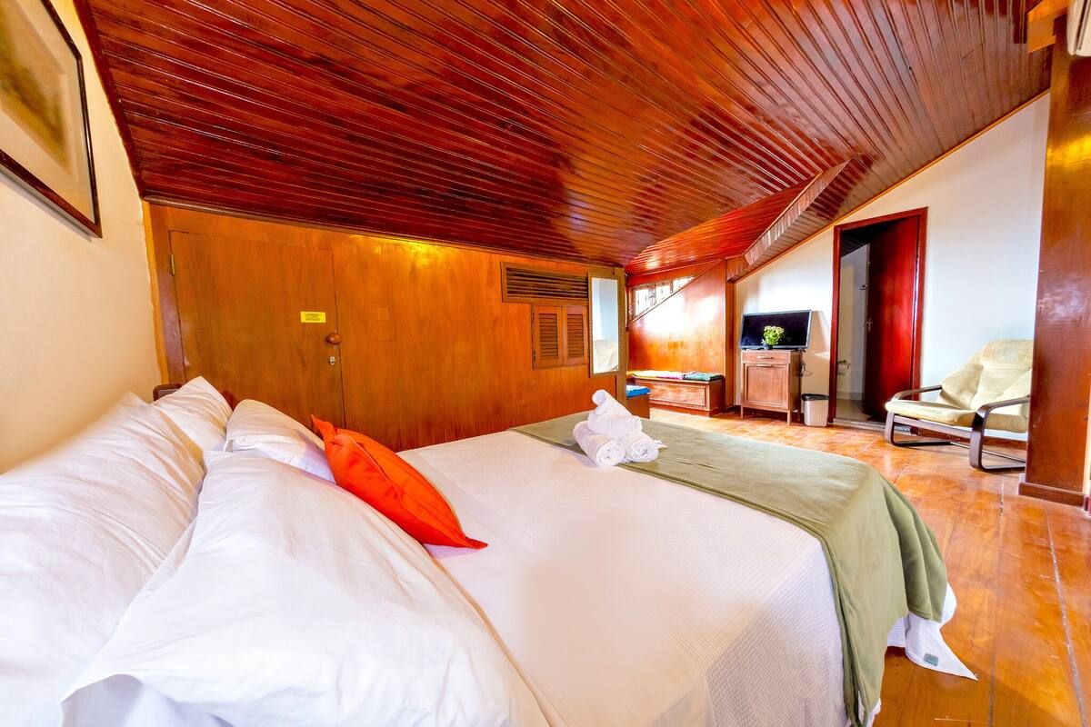A hotel room with a wooden ceiling, a bed with white linens, a green throw blanket, and a chair.