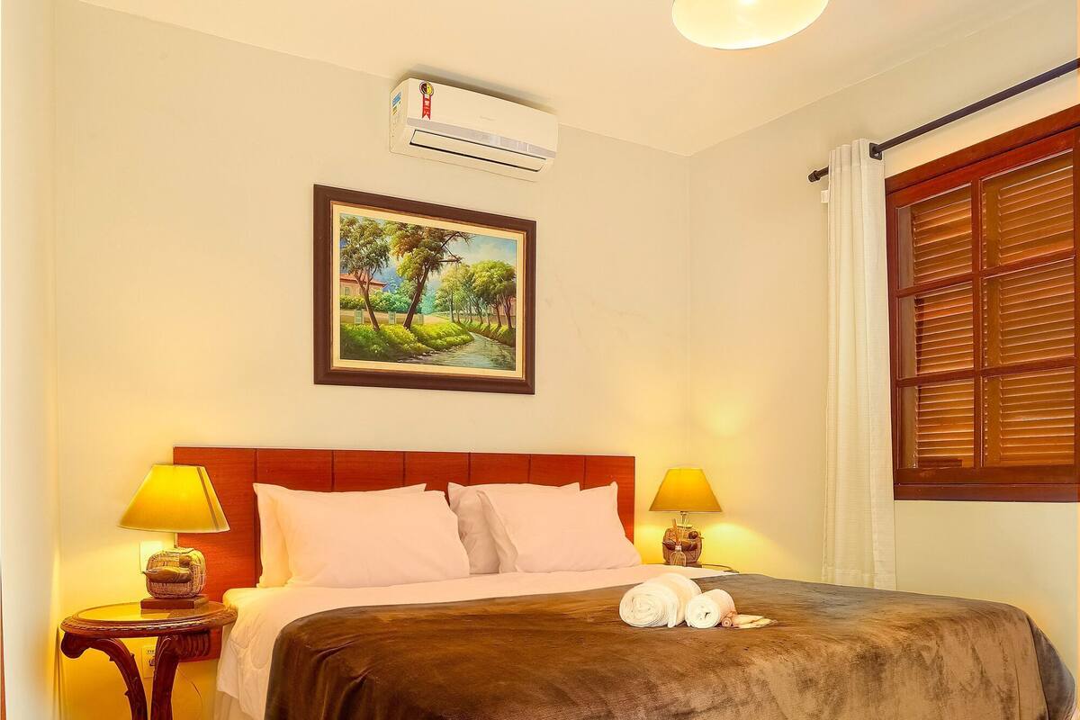 A neatly made bed with a brown blanket, two bedside lamps, a framed picture on the wall, and an air conditioning unit above the bed.