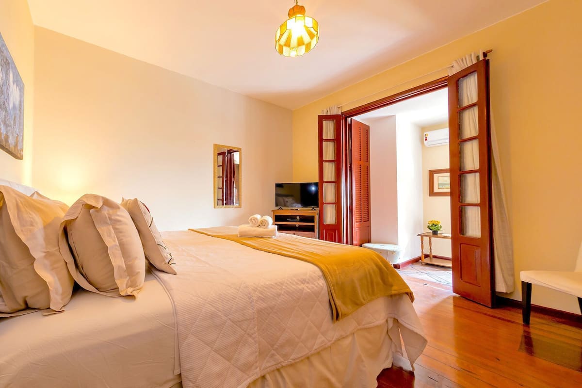 A bedroom with a bed, a television, a wooden floor, and a door leading to another room.
