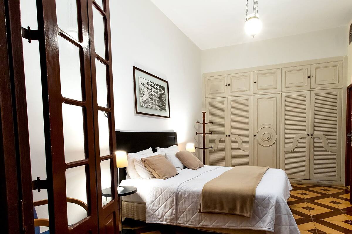A bedroom with a bed, a nightstand, a wall-mounted lamp, a wardrobe, and a geometric patterned floor.