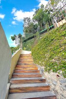 Uma escadaria de madeira que sobe por uma parede de pedra, cercada por vegetação e um céu azul límpido.