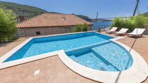 Une piscine avec une terrasse carrelée, un espace détente avec des fauteuils et une vue sur un pont et des collines en arrière-plan.