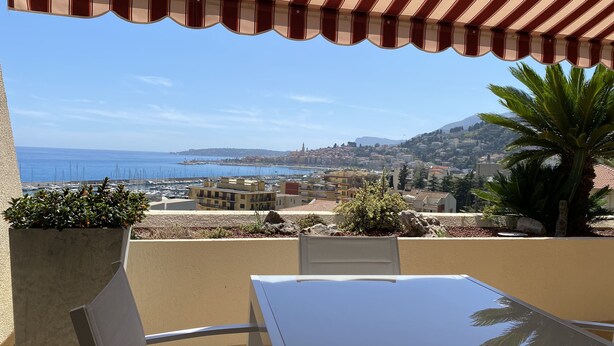 Ein Balkon mit gestreiftem Sonnenschirm, einem Tisch, Stühlen und Blick auf das Meer und die Stadt.
