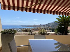 Un balcó amb una estoreta a ratlles, una taula, cadires i vistes al mar i al paisatge urbà.
