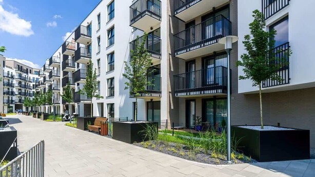 A modern apartment building with multiple balconies, a paved walkway, and small trees.