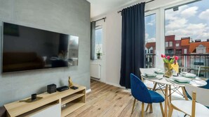 A modern living room with a flat-screen TV, wooden flooring, a dining area with a table set for two, and large windows with curtains.