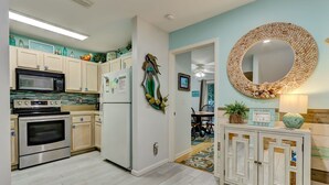 Una cocina con armarios blancos, horno de acero inoxidable, microondas y refrigerador. Hay un espejo decorativo en la pared y un mueble con espejo.
