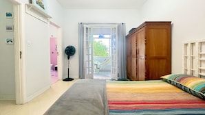 Une chambre avec un lit, une armoire en bois, un ventilateur sur pied et une vue sur un balcon, accessible par une porte ouverte.