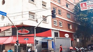 Une vue de rue avec un immeuble doté de nombreuses fenêtres et d’une façade en briques rouges. On y voit une enseigne pour « Bia Lanches » et un panneau indiquant un passage piéton.