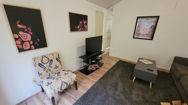 Ein Wohnzimmer mit einem gemusterten Sessel, einem Flachbildfernseher auf einem schwarzen Standfuß, einem grauen Sofa und einem Bilderrahmen mit Blumen an der Wand.
