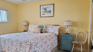 A bedroom with a bed, two bedside tables, a chair, a ceiling fan, and a beach-themed picture on the wall. - Private Beach Living (Cocoa Beach)