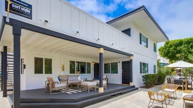 Una casa de dos pisos con un patio cubierto, mobiliario exterior y un letrero que anuncia 'Surf Style Vacation Homes'.