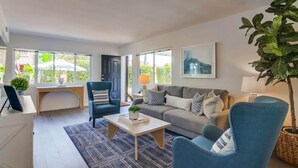 Una sala de estar moderna con sofá, sillones, una mesa de centro y un escritorio. Amplias ventanas ofrecen luz natural y vistas al exterior.