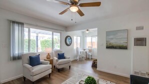 Una sala de estar amplia con un ventilador de techo, dos sillones blancos, una mesa de centro y un comedor con mesa y sillas.