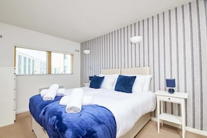 A neatly made bed with blue and white bedding, a bedside table with a lamp, and a window with blinds. - LS1 Perfect For Business Travellers & Relocations - 4 Beds, 2 Baths - FREE WiFi (Leeds)