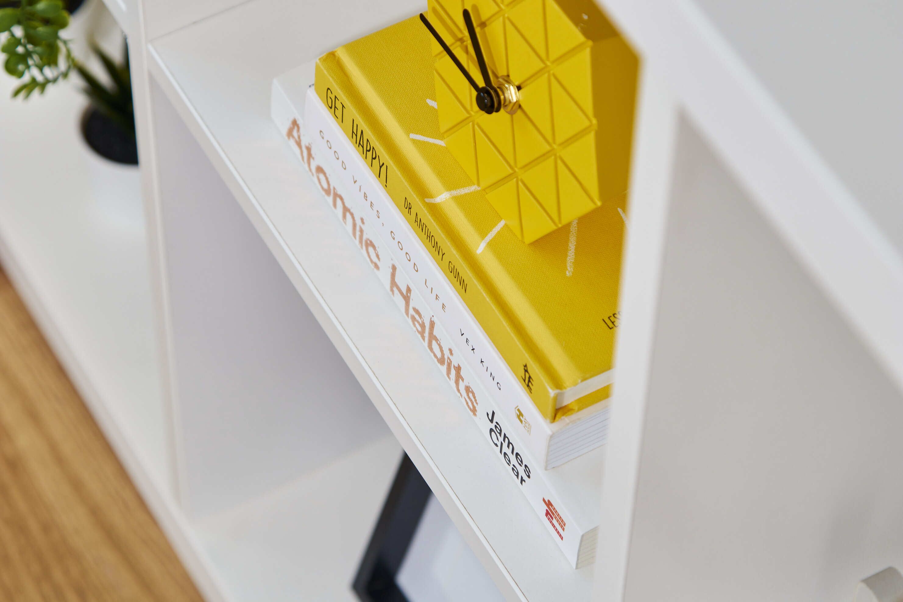 Una estantería blanca con un libro titulado 'Good Vibes, Good Life' y una caja de regalo amarilla con un reloj encima.