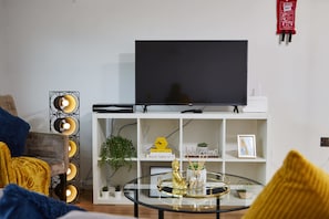 A living room with a flat-screen TV on a white stand, a glass coffee table with decorative items, a bookshelf with books and a welcome sign, and a floor lamp with multiple bulbs. - LS1 Perfect For Business Travellers & Relocations - 4 Beds, 2 Baths - FREE WiFi (Leeds)
