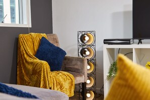 A room with a chair covered in a yellow blanket, a blue pillow, and a floor lamp with multiple bulbs. - LS1 Perfect For Business Travellers & Relocations - 4 Beds, 2 Baths - FREE WiFi (Leeds)