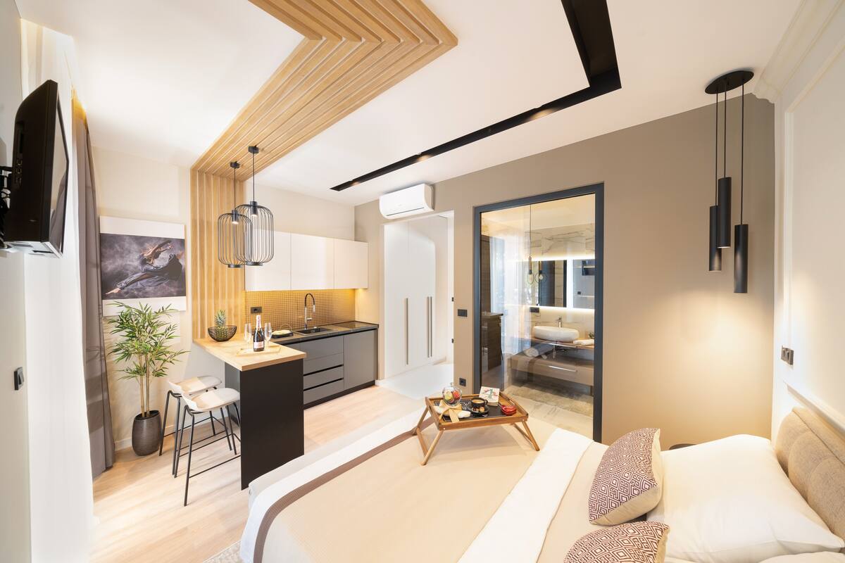 A modern hotel room with a large bed, a small kitchenette, a dining area, and a bathroom.