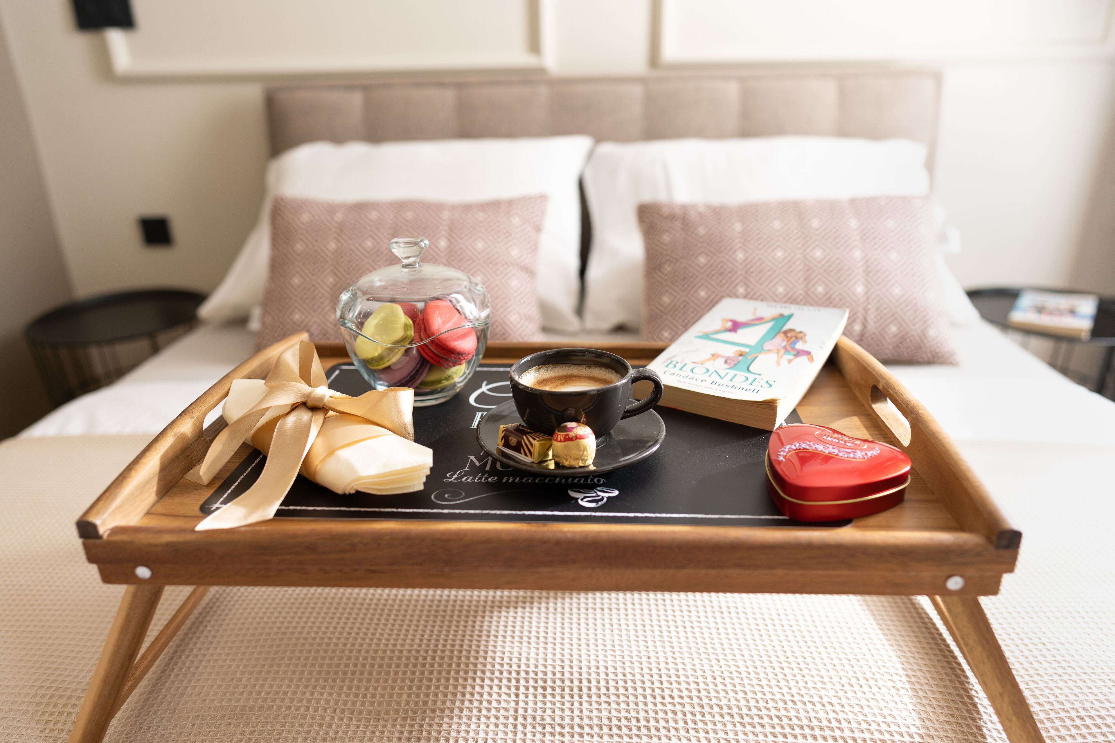 Un plateau de petit-déjeuner avec une tasse de café, des macarons, un livre et un récipient en forme de cœur, posé sur un lit.