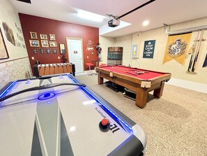 A pool table, foosball table, and a billiards table in a room with a brick wall and framed pictures.
