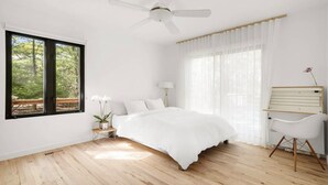 A bedroom with a bed, a ceiling fan, a bedside table with a lamp, a chair, and a window with a view of trees. - Modern Newly Renovated Summer Get Away  (East Hampton)