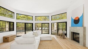A modern living room with a white sofa, a fireplace, and large windows offering a view of the outdoors. - Modern Newly Renovated Summer Get Away  (East Hampton)