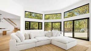 A modern living room with a white sectional sofa, a large window with a forest view, and a staircase leading to another level. - Modern Newly Renovated Summer Get Away  (East Hampton)