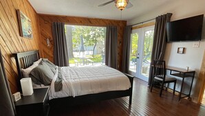 Une chambre à coucher avec un lit, un bureau et une chaise. Il y a une fenêtre avec des rideaux, un ventilateur de plafond et un téléviseur à écran plat fixé au mur.