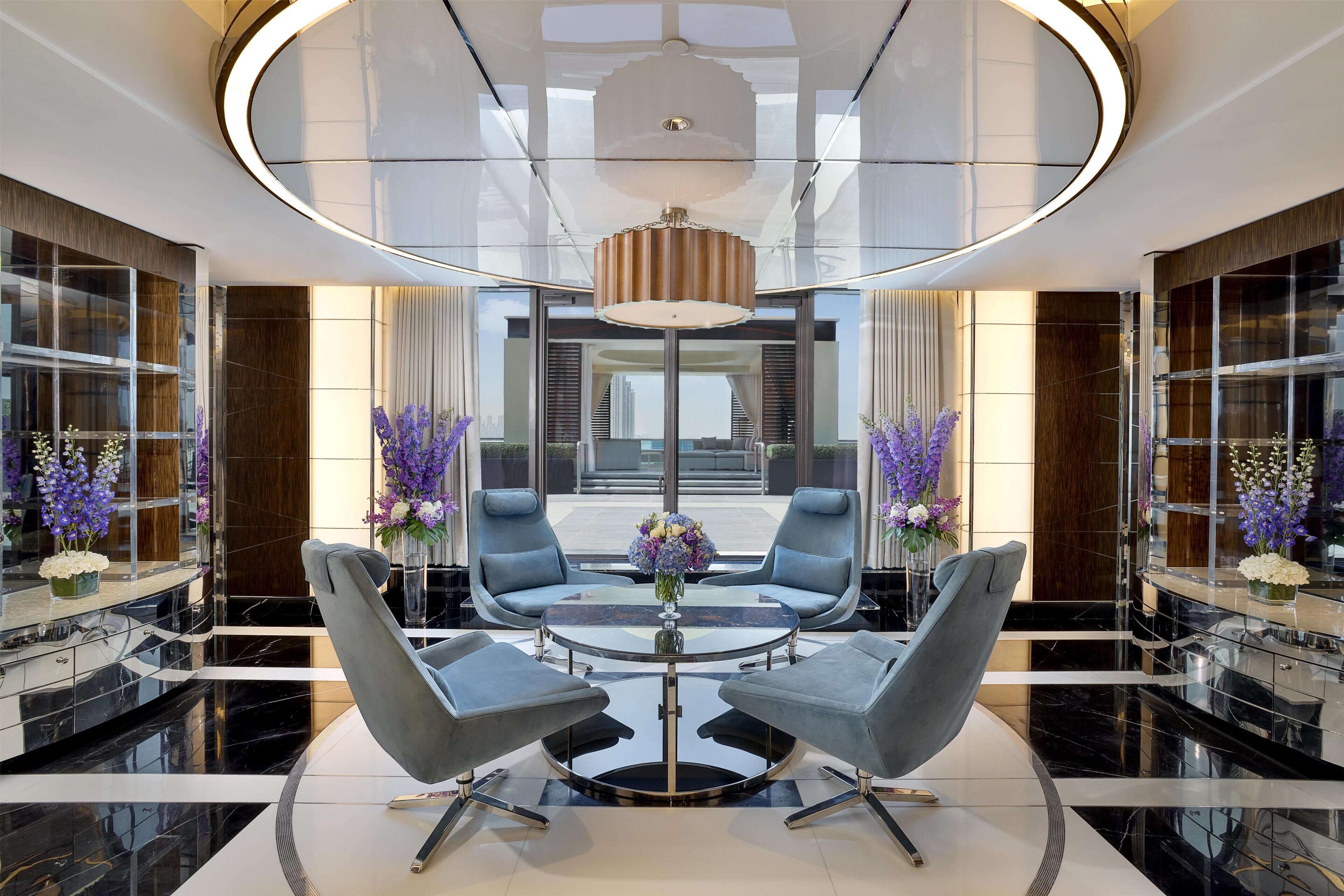 Un hall d’entrée moderne avec une table basse en verre entourée de fauteuils gris, un lustre central et des fleurs violettes.