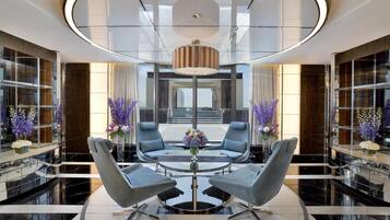 Un hall d’entrée moderne avec une table basse en verre entourée de fauteuils gris, un lustre central et des fleurs violettes.