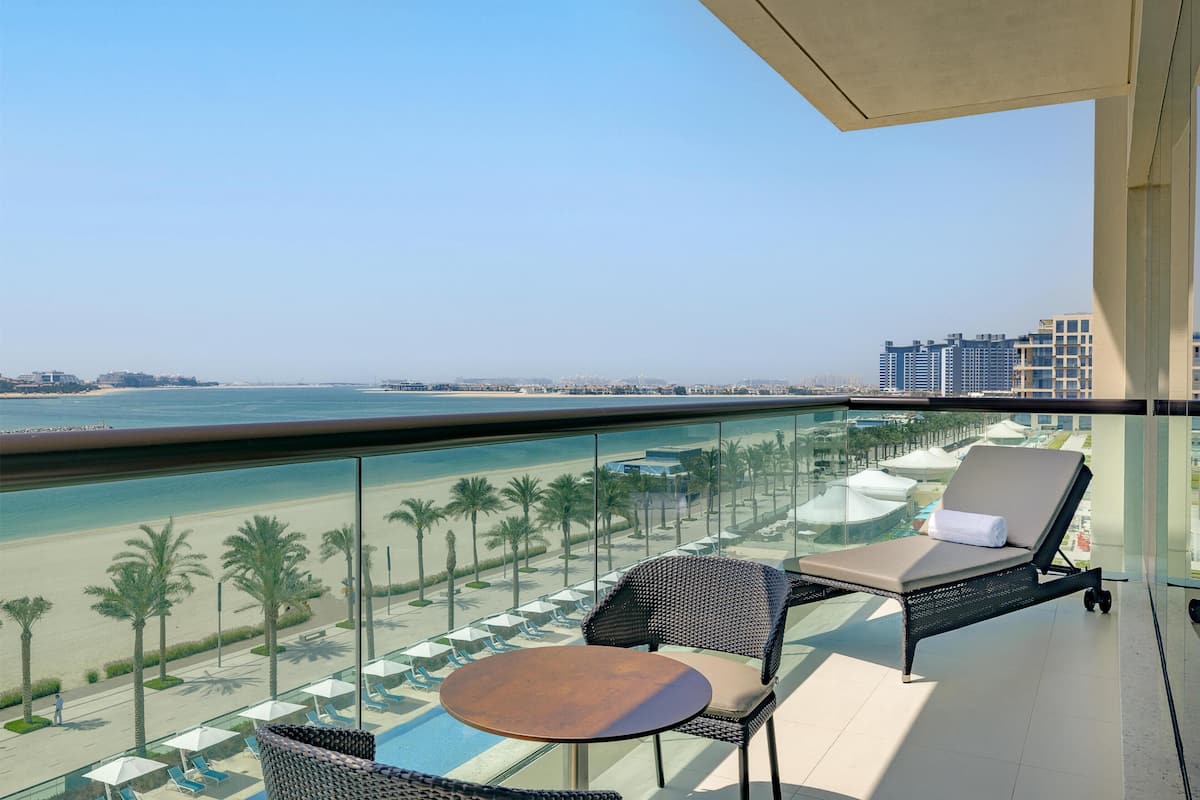 Ein Balkon mit Glasgeländer, der auf einen Strand blickt, mit Liegestühlen und einem kleinen Tisch.