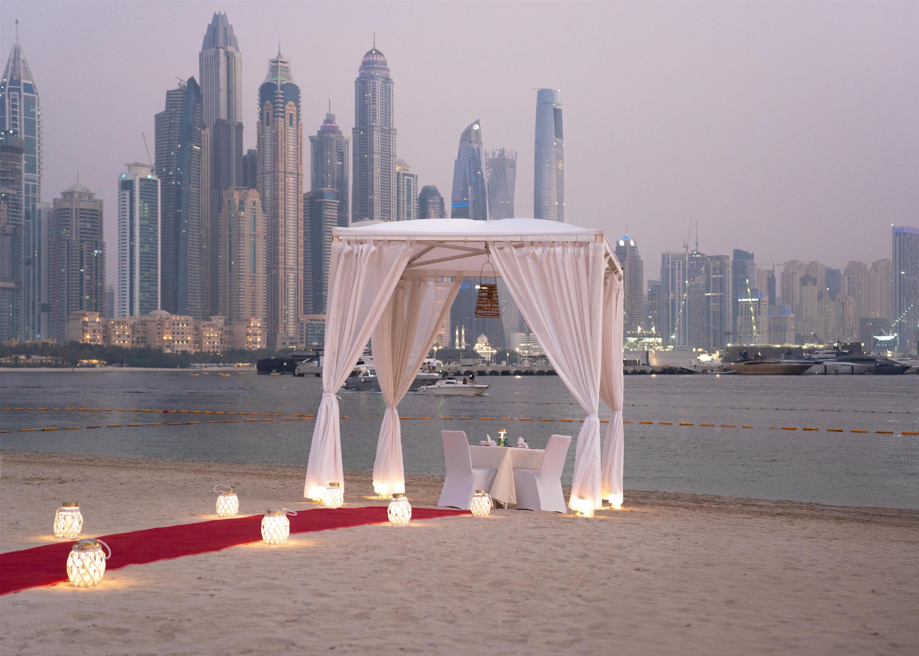 Une cérémonie de mariage organisée sur la plage, avec un auvent et des tables, offrant une vue sur les gratte-ciel d’une ville.