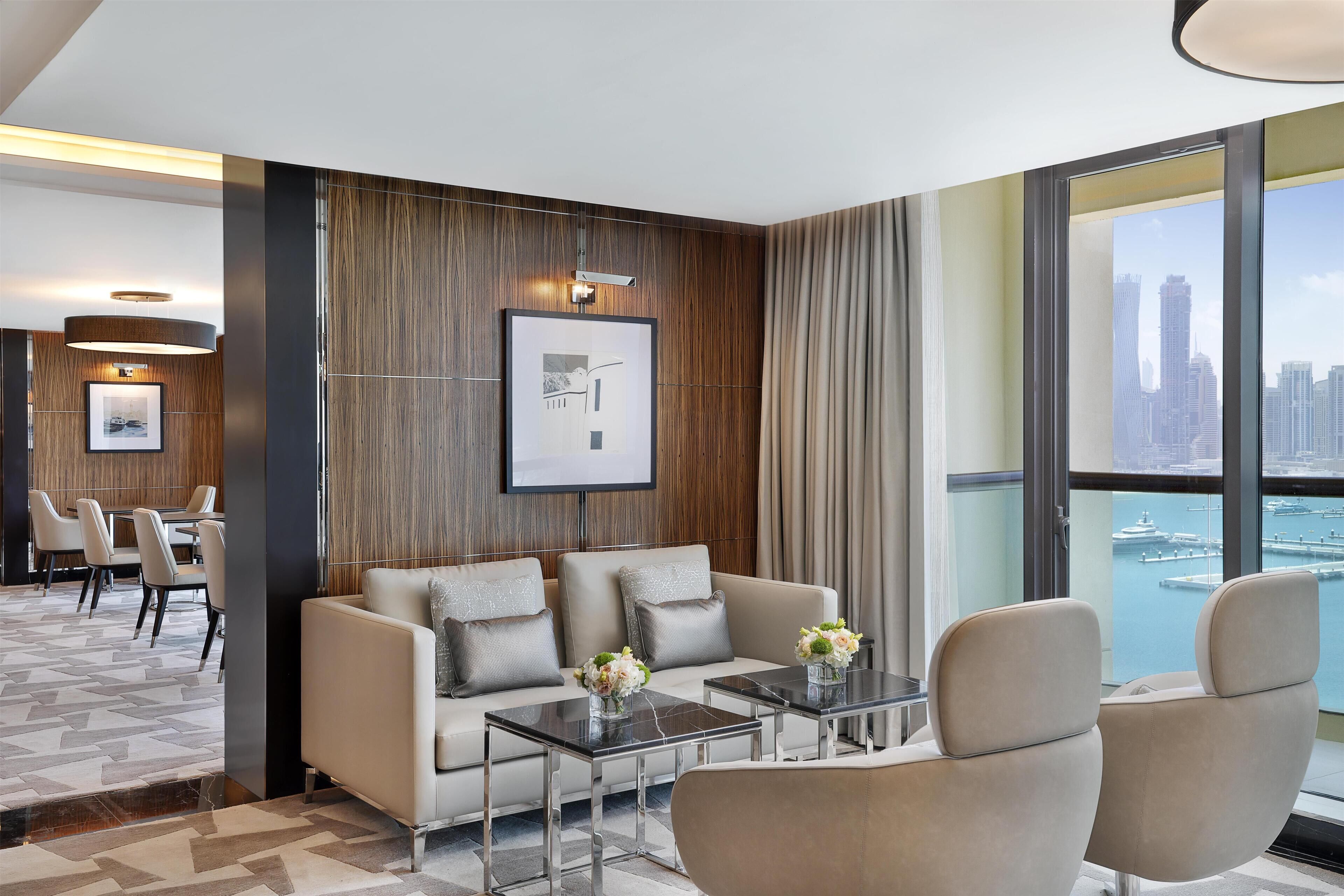 A modern hotel room with a sofa, two armchairs, a coffee table, and a view of a cityscape and water.