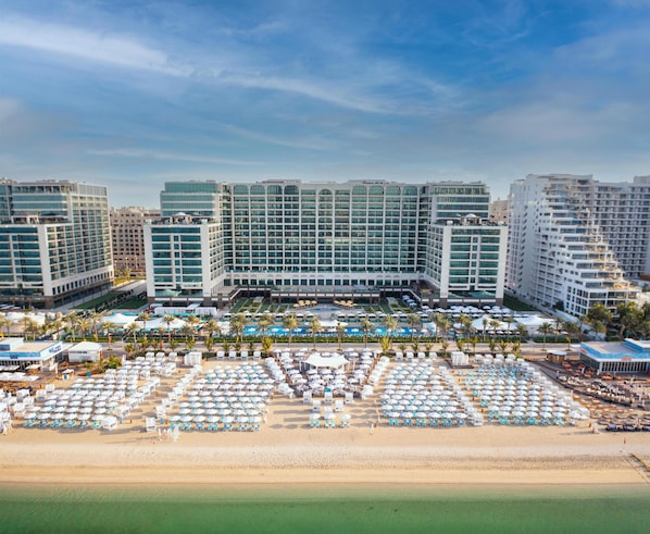 A hotel with multiple floors, a beach area with numerous umbrellas and sun loungers, and a clear view of the ocean.