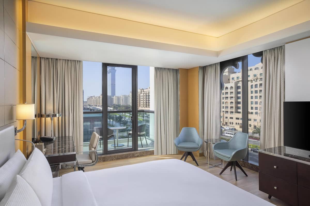 Ein modernes Hotelzimmer mit einem großen Bett, einem Schreibtisch mit Lampe, zwei Stühlen, einem Fernseher und einem Balkon mit Blick auf die Stadt.