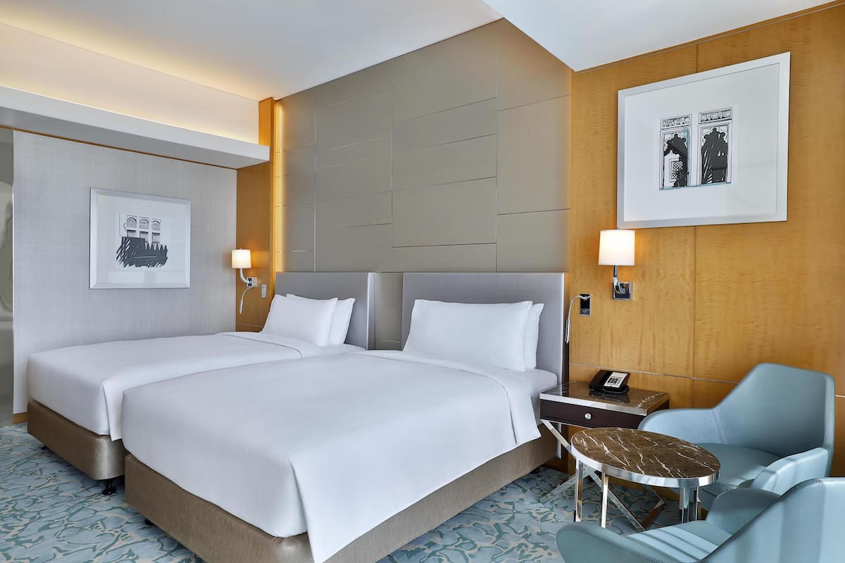 Ein Hotelzimmer mit zwei Betten, einem Schreibtisch mit Telefon, einem Sessel und einem kleinen Tisch.
