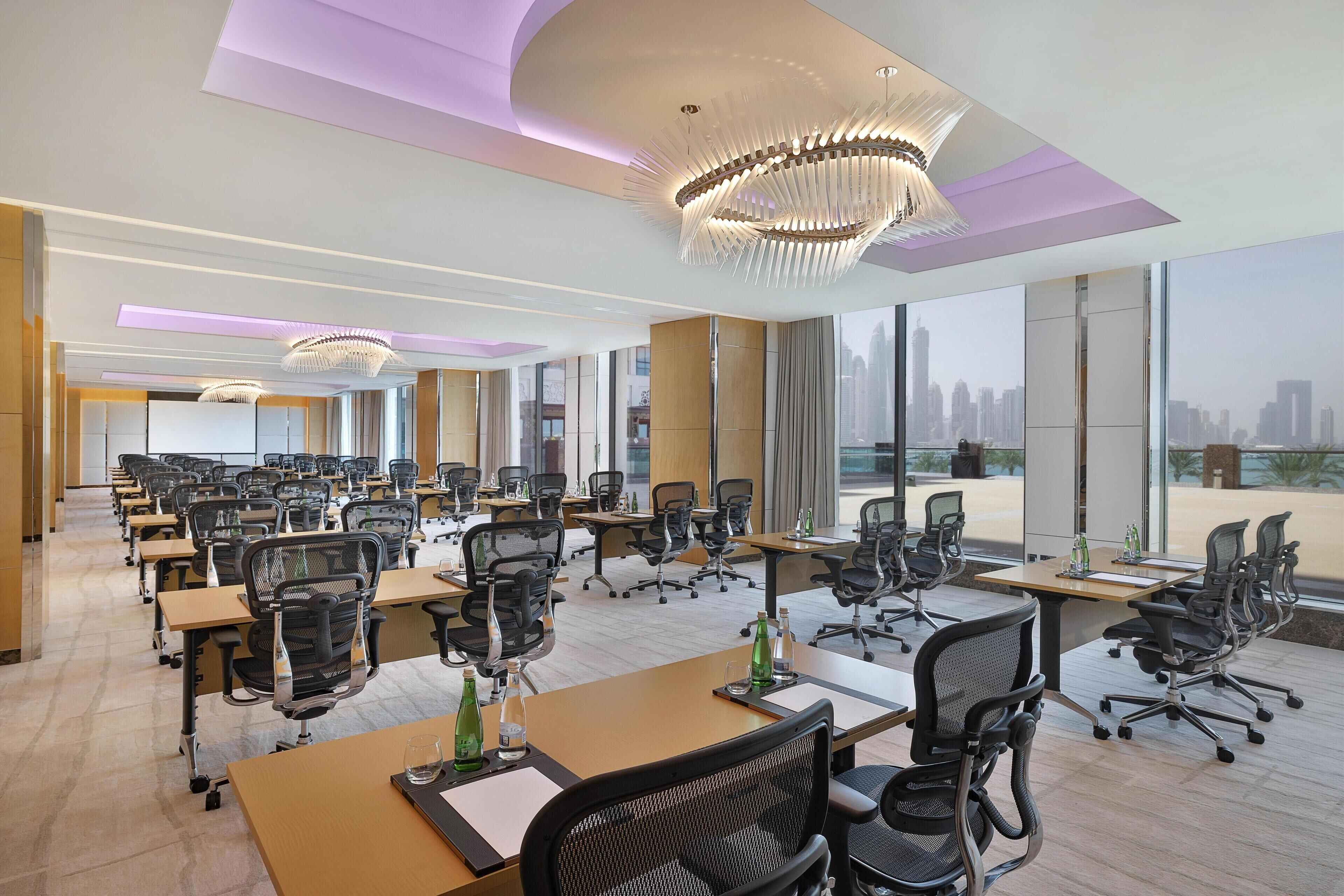 A modern conference room with multiple tables and chairs, large windows with a city view, and contemporary lighting fixtures.