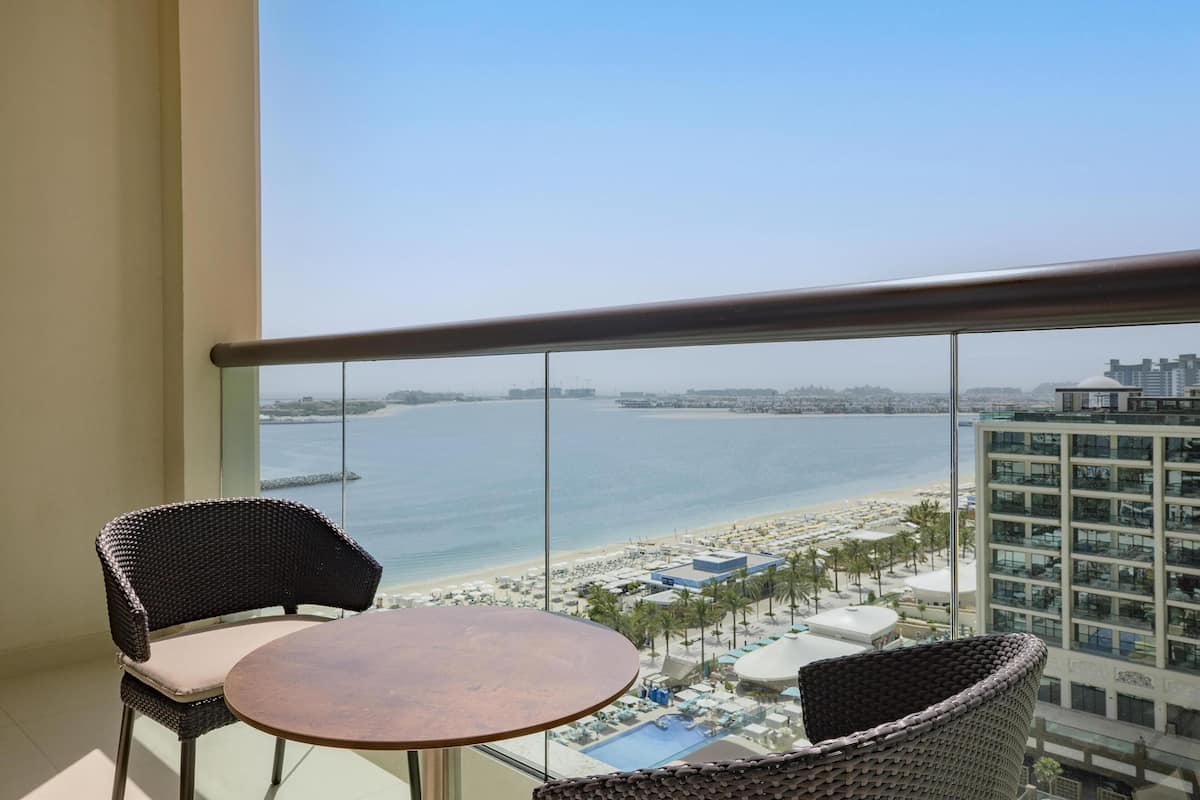 Ein Balkon mit Korbstühlen und einem kleinen Tisch, mit Blick auf einen Strand und einen Poolbereich.