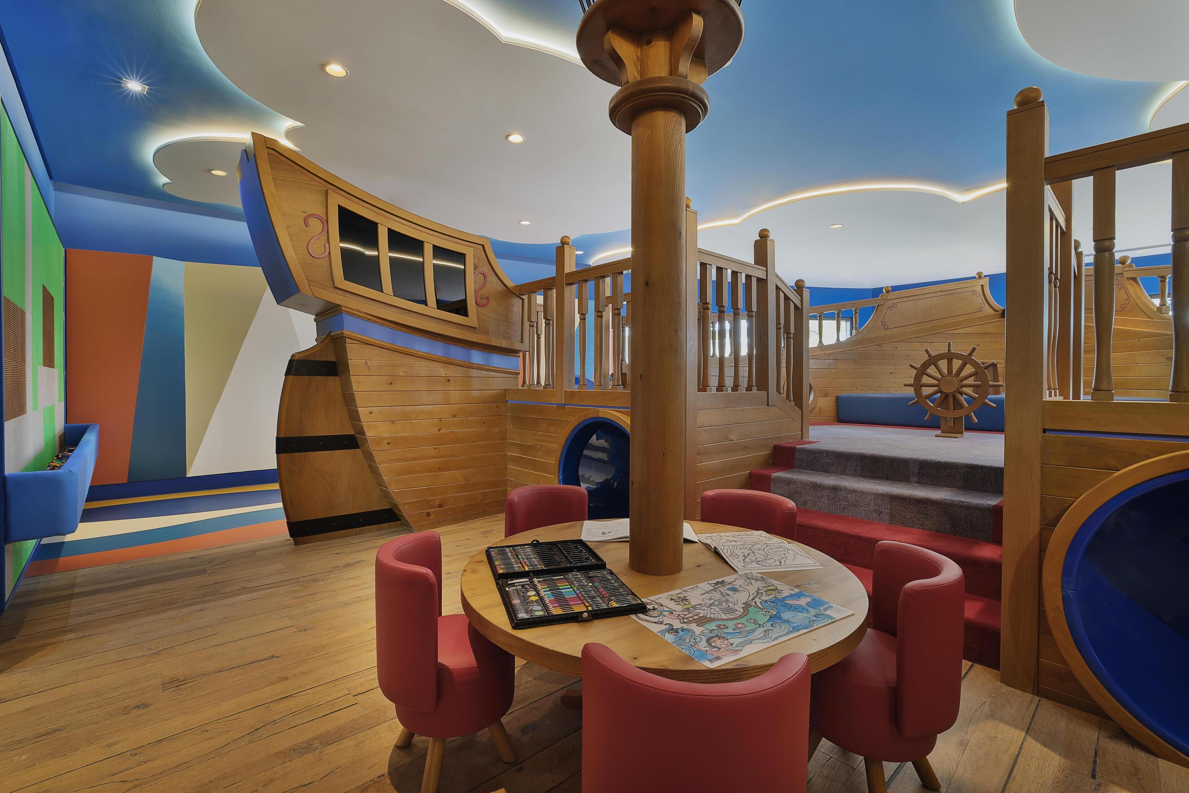 A play area with a wooden ship-themed play structure, a round table with chairs, and a slide.