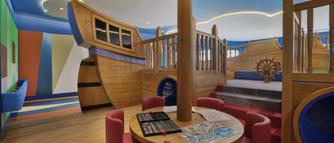 A play area with a wooden ship-themed play structure, a round table with chairs, and a slide.