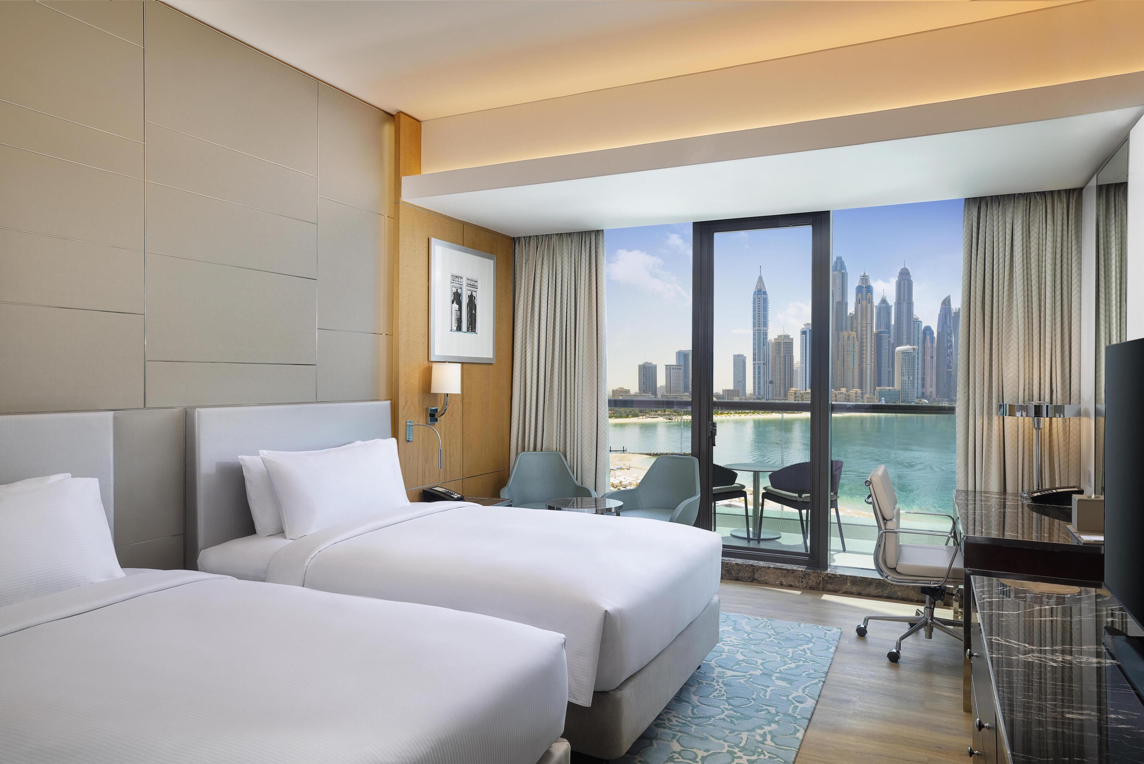 A hotel room with two beds, a desk, a chair, a TV, and a large window with a cityscape view.